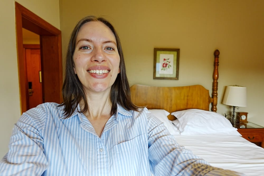 Audrey Bergner smiles for a selfie inside a Llao Llao Hotel room in Bariloche, Río Negro, Patagonia, Argentina, with a carved wooden bed, bedside table and framed floral artwork behind her.
