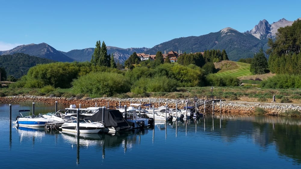 Boats moored at the Llao Llao marina in Bariloche, Río Negro, Argentina, with calm lake water, the historic hotel on a wooded rise and Patagonian mountains beneath a clear blue sky.