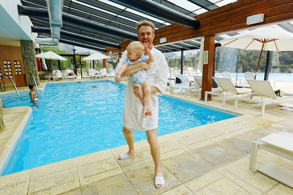 Samuel Jeffery carries toddler Aurelia Jeffery beside the indoor pool at Llao Llao Resort in Bariloche, Río Negro, Argentina, with glass walls, timber beams, loungers and the outdoor pool beyond.