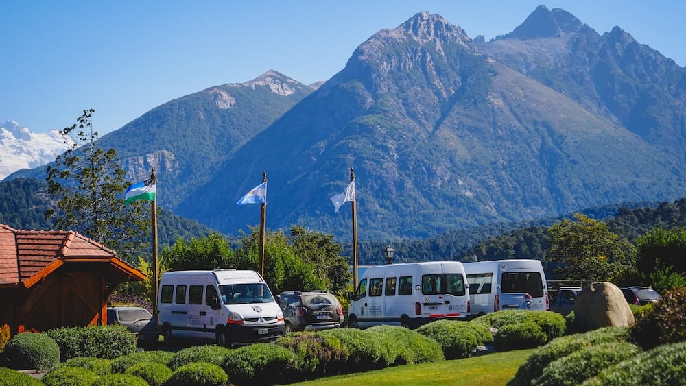 Shuttle vans and parked vehicles outside Llao Llao Resort in Bariloche, Río Negro, Argentina, framed by manicured gardens, flagpoles and steep Patagonian mountain peaks under a clear blue sky.