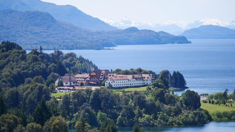 Distant view of Llao Llao Resort in San Carlos de Bariloche, Río Negro, Argentina, surrounded by forest beside Nahuel Huapi Lake with layered Patagonian mountains and snowy peaks beyond.