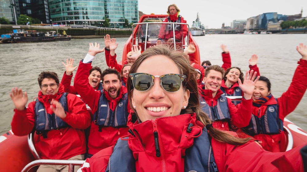 London, England travel campaign scene on the River Thames, with a group of creators including Audrey Bergner in red jackets waving on a speedboat, capturing a high-energy tourism project in the city’s waterfront district.
