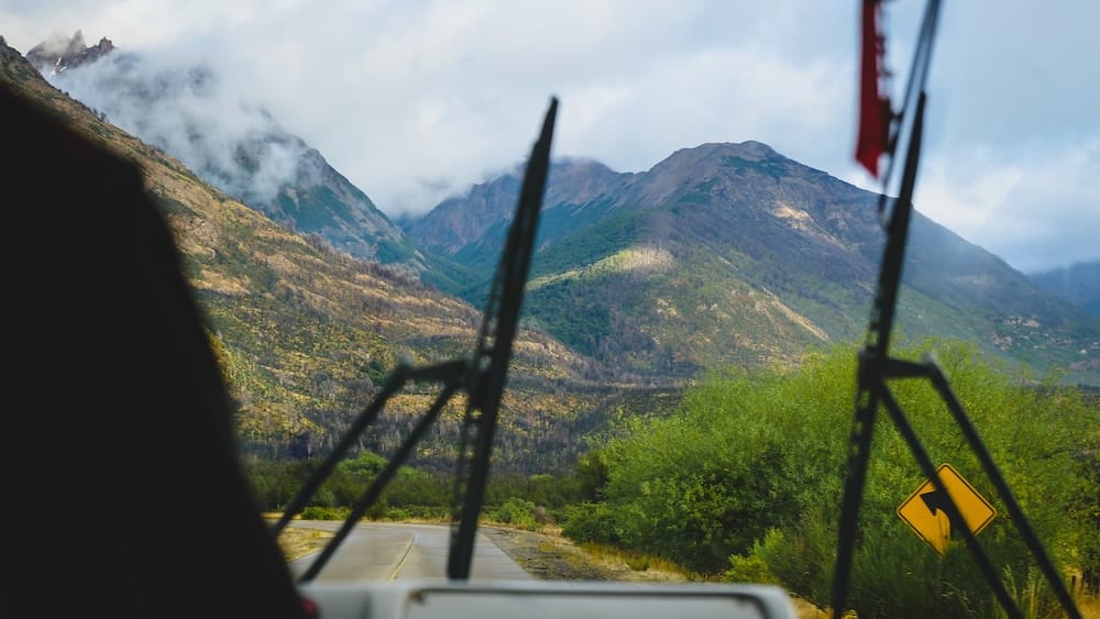 View from a bus traveling through Los Alerces National Park in Patagonia Argentina, showing a winding road, dense forest, and Andean mountains under shifting clouds.

