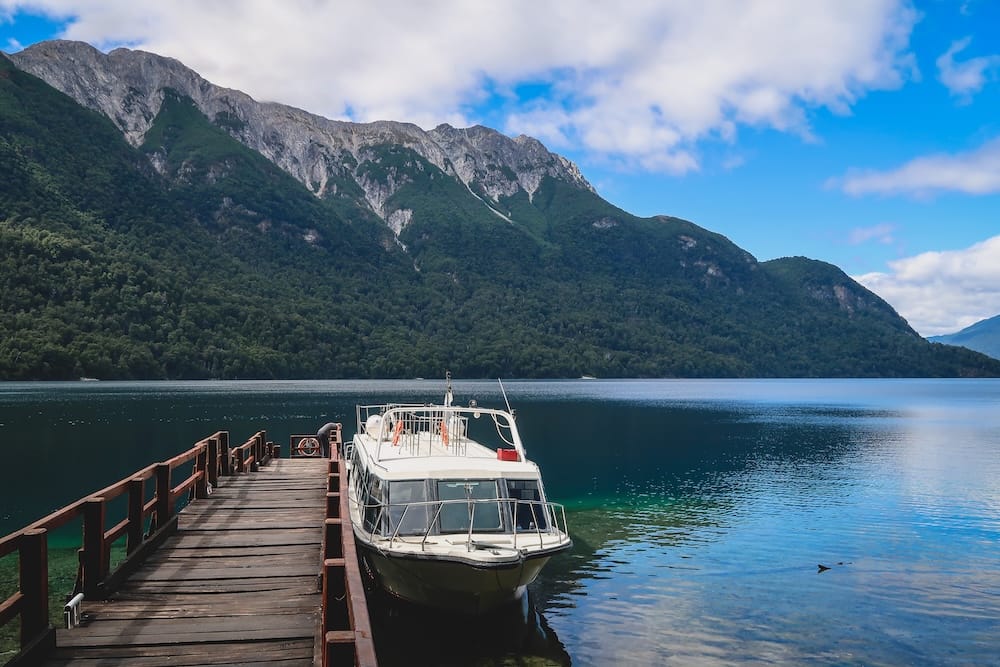 A small excursion boat is moored beside a wooden pier in Los Alerces National Park, Chubut, Patagonia, Argentina, with clear lake water, dense forest and steep rocky mountains rising behind it.