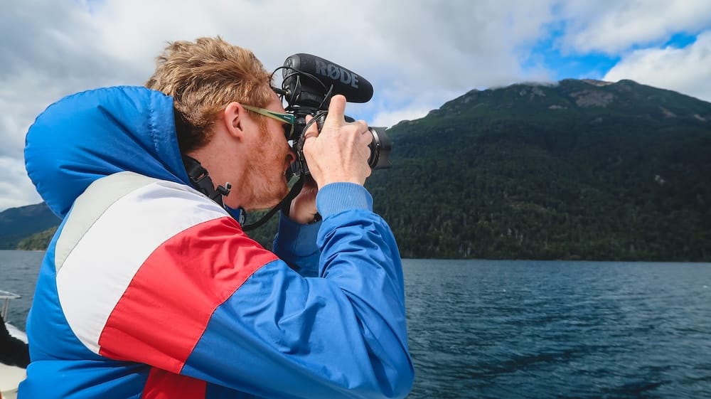 Samuel Jeffery filming mountain lake scenery in Los Alerces National Park, Chubut, Patagonia, Argentina Samuel Jeffery films from a boat on a deep blue mountain lake in Los Alerces National Park, with forested slopes and cloud-covered peaks rising across the water in Chubut, Patagonia, Argentina.