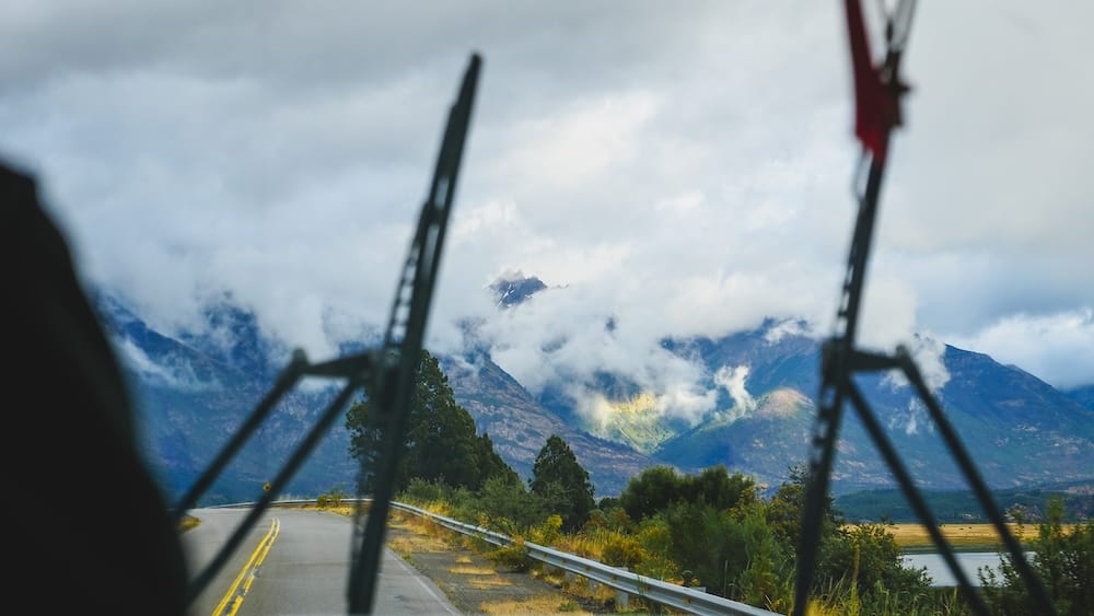 Cloud-covered Andean peaks rise above a winding paved road in Los Alerces National Park, Chubut, Patagonia, Argentina, viewed through a vehicle windshield with wipers framing the mountain landscape.