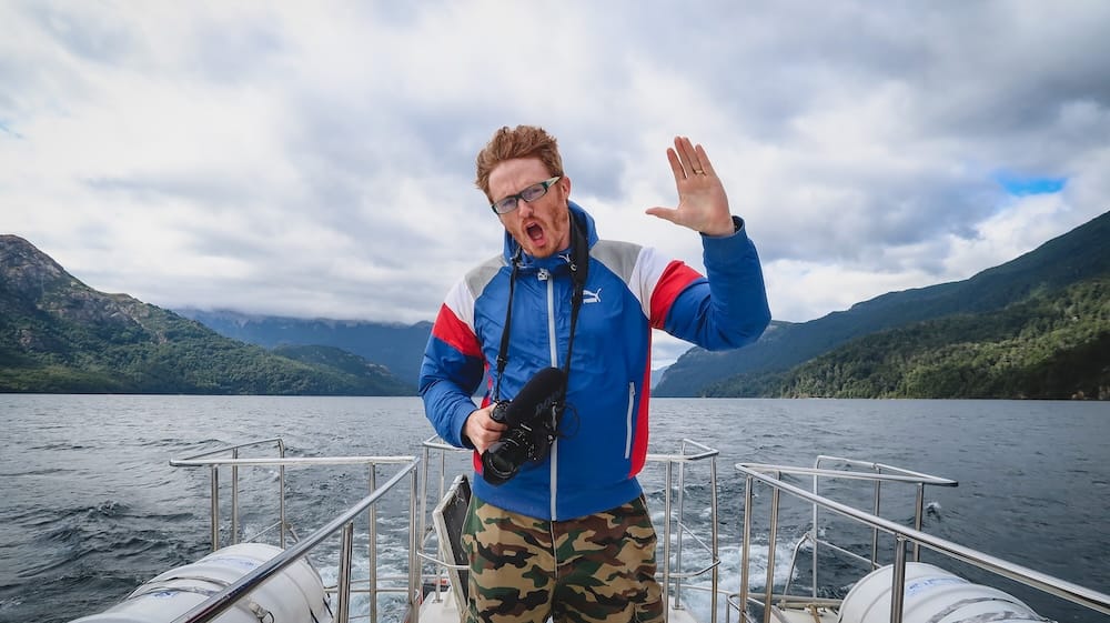 Samuel Jeffery stands on a boat in Los Alerces National Park, Patagonia, Argentina, holding a camera and reacting to strong wind above the deep glacial lake with forested mountains behind him.