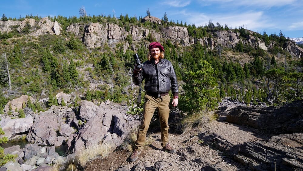 Samuel Jeffery photographing rugged terrain in Los Alerces National Park near Trevelin Patagonia Argentina Samuel Jeffery standing on rocky cliffs in Los Alerces National Park near Trevelin Chubut Argentina during autumn, holding a camera and overlooking rugged Patagonian terrain with forested foothills under clear skies.