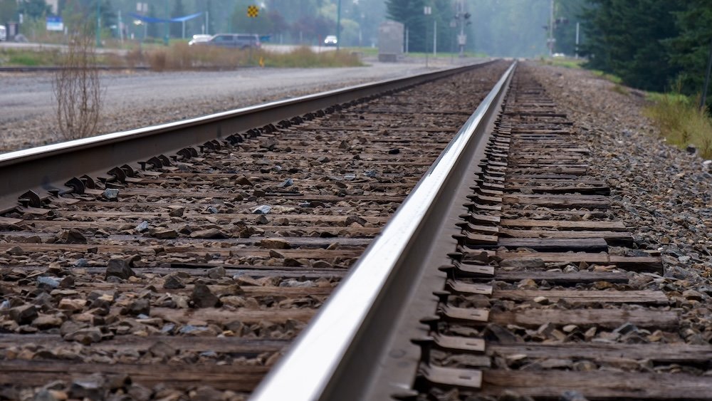 Low-angle railway track view in Fernie BC A low-angle view of railway tracks in Fernie, British Columbia, highlighting the wooden ties, gravel ballast, and long straight rail stretching into the distance, with soft morning haze and hints of traffic and forested surroundings in the background.