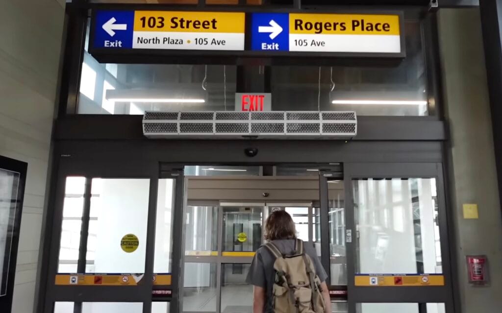 Salida del LRT hacia Rogers Place en el centro de Edmonton, Alberta, Canadá, con Samuel Jeffery Samuel Jeffery camina hacia la salida de una estación del LRT de Edmonton bajo carteles que indican Rogers Place y 103 Street, en el centro de Edmonton, Alberta, Canadá, con puertas de vidrio y señalización al frente.