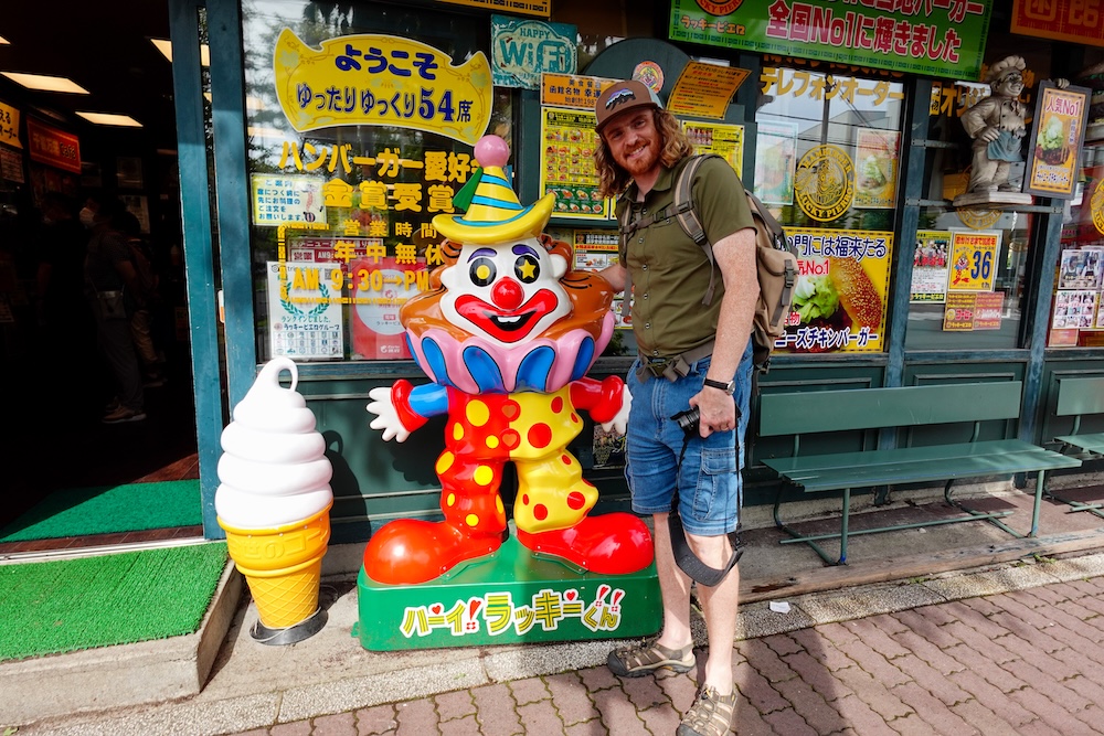 Samuel Jeffery sonríe junto a una colorida mascota de payaso de Lucky Pierrot afuera de la hamburguesería Goryokaku Park Front en Hakodate, Hokkaido, Japón, con carteles de menú en japonés y un gran cono de helado decorativo.