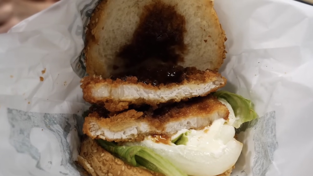 Primer plano de una hamburguesa de tonkatsu de cerdo en Lucky Pierrot en Hakodate, Hokkaido, Japón, cortada al medio para mostrar dos piezas de cerdo rebozado, salsa oscura, lechuga y mayonesa dentro de un pan con sésamo y envoltorio de papel blanco.