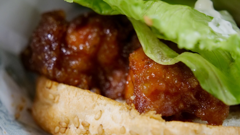 Primerísimo plano de la Chinese Chicken Burger en Lucky Pierrot en Hakodate, Hokkaido, Japón, mostrando trozos brillantes de pollo frito con glaseado dulce debajo de lechuga verde crocante y mayonesa dentro de un pan con semillas de sésamo.