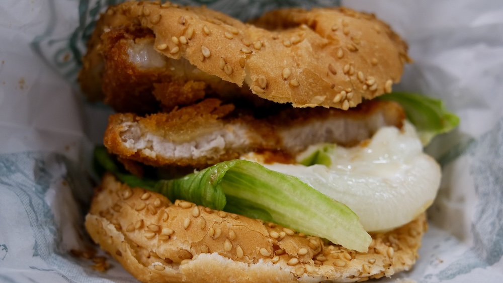 Hamburguesa tonkatsu de Lucky Pierrot con cerdo empanado, lechuga y mayonesa en pan con sésamo, Hakodate, Hokkaido, Japón Primer plano de una hamburguesa tonkatsu de Lucky Pierrot en Hakodate, Hokkaido, Japón, con una milanesa de cerdo empanada, lechuga verde y mayonesa cremosa entre panes con semillas de sésamo, parcialmente envuelta en papel.