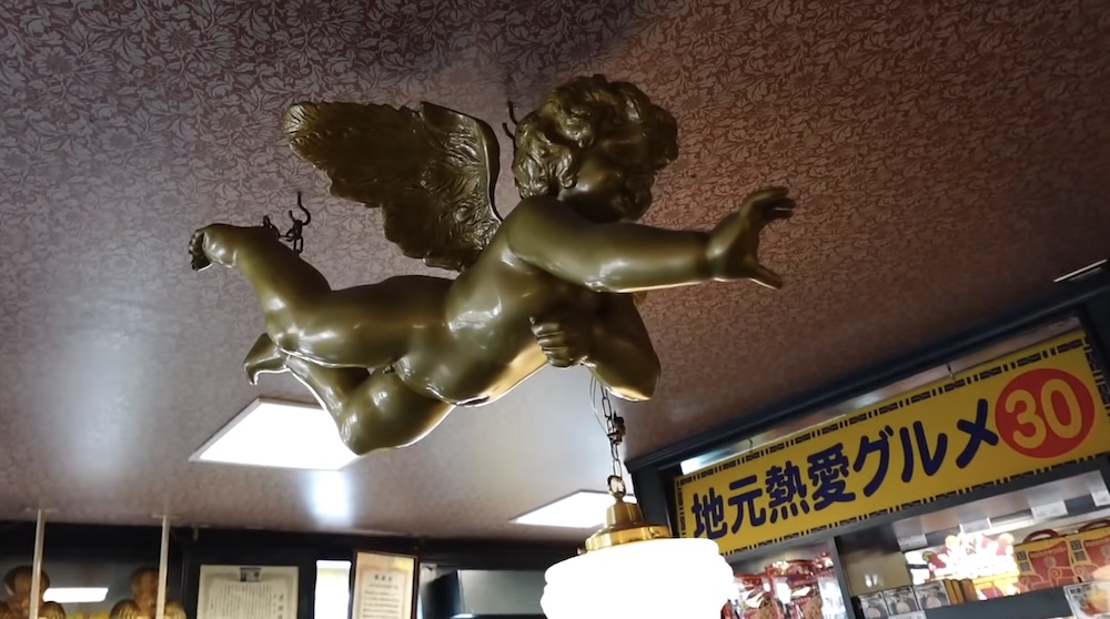 Querubín colgante en el interior de Angels’ Chatter, Lucky Pierrot cerca de Goryokaku, Hakodate, Hokkaido, Japón Estatua de querubín alado color bronce colgada de un techo estampado dentro de un restaurante Lucky Pierrot en Hakodate, Hokkaido, Japón, junto a luces de techo y un cartel amarillo en japonés.