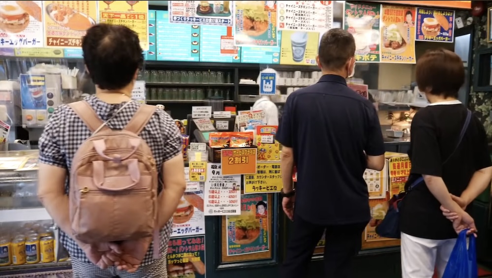 Mostrador de pedidos y carteles del menú en Lucky Pierrot cerca de la Torre Goryokaku, Hakodate, Hokkaido, Japón Clientes de espaldas a la cámara en el mostrador de un restaurante Lucky Pierrot en Hakodate, Hokkaido, Japón, frente a carteles de menú en japonés llenos de fotos de hamburguesas y avisos.