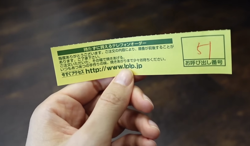 Ticket de pedido numerado en la mano de Audrey Bergner en Lucky Pierrot cerca de Goryokaku, Hakodate, Hokkaido, Japón Mano de Audrey Bergner sosteniendo un ticket de pedido amarillo de Lucky Pierrot impreso en japonés con un número escrito a mano en un recuadro, sobre una mesa de madera oscura en Hakodate, Hokkaido, Japón.