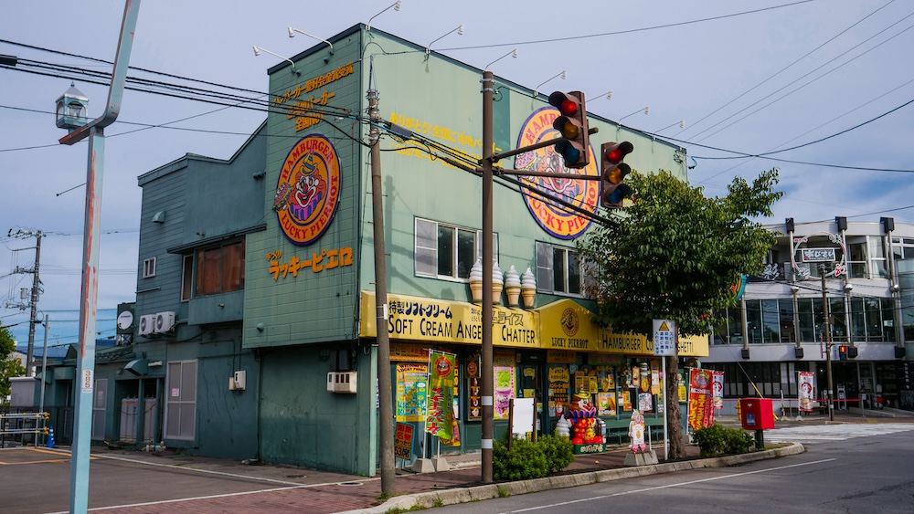 Exterior en esquina de la sucursal de Lucky Pierrot cerca de la Torre Goryokaku, Hakodate, Hokkaido, Japón Restaurante Lucky Pierrot verde de dos pisos en una esquina de Hakodate, Hokkaido, Japón, con logos redondos de payaso, conos de helado en el techo, cartelería amarilla sobre la entrada y semáforos al frente.