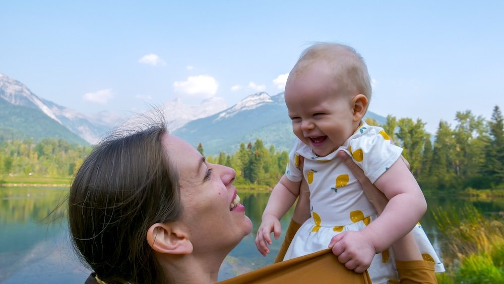 Audrey and baby Aurelia at Maiden Lake in Fernie Audrey Bergner of That Backpacker holds baby Aurelia at Maiden Lake in Fernie, British Columbia, sharing a joyful moment with mountain peaks and a calm reflective lake behind them, capturing a family-friendly stop on this scenic trail.
