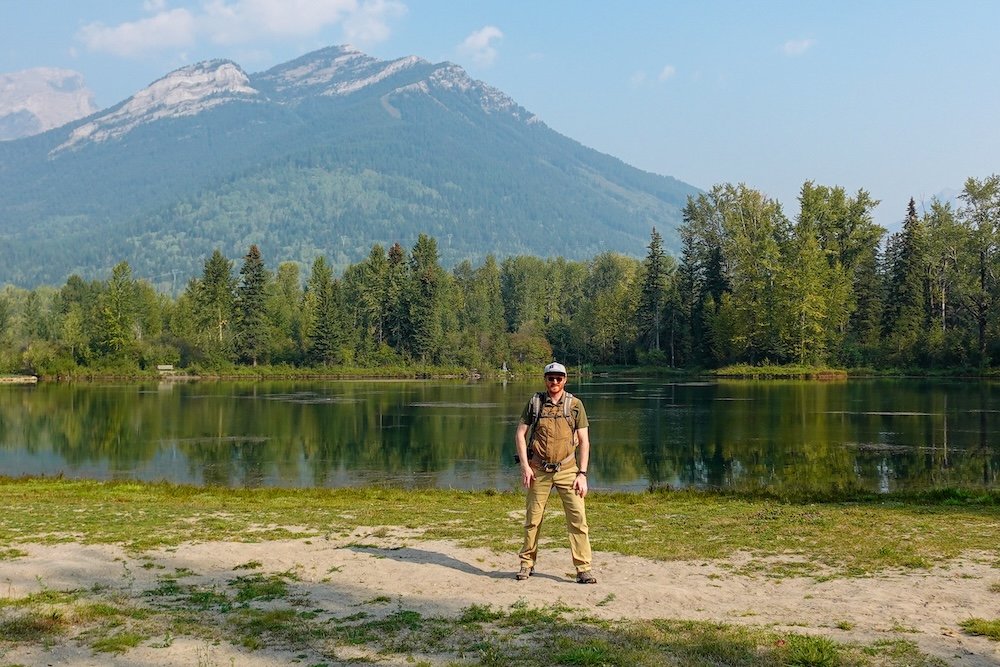 Maiden Lake mountain views in Fernie Nomadic Samuel Jeffery standing beside Maiden Lake in Fernie, British Columbia, with calm water reflections, forested shoreline, and mountain peaks in the background, capturing an easy outdoor moment during a relaxed summer walk near downtown.