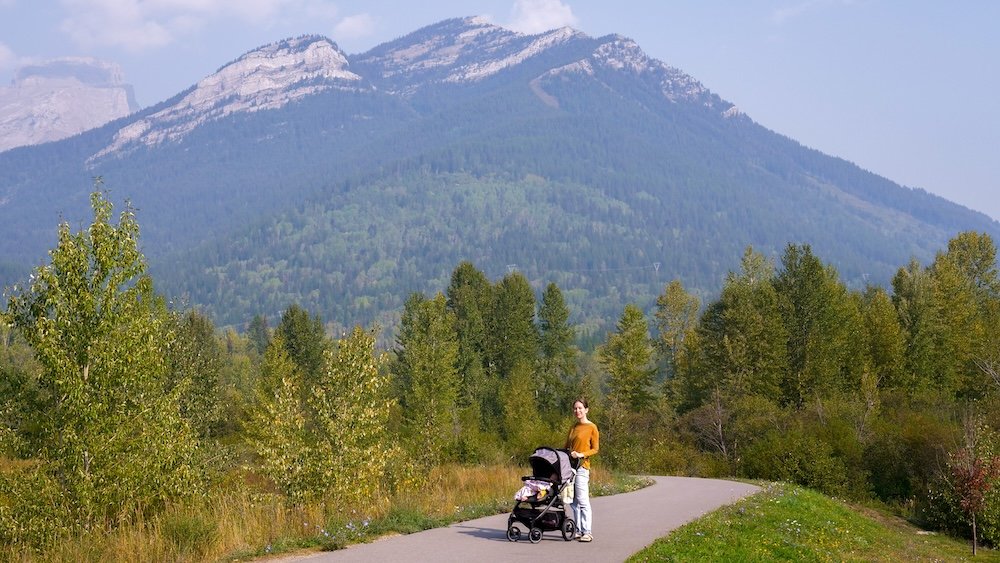 Audrey and baby Aurelia walking at Maiden Lake in Fernie Audrey Bergner of That Backpacker walks with baby Aurelia in a stroller along the paved trail at Maiden Lake in Fernie, British Columbia, surrounded by lush trees and framed by the dramatic peaks of the Lizard Range on a calm late-summer day.