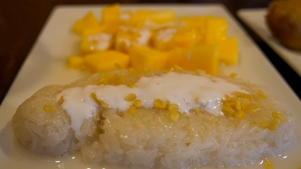 Macro close-up of mango sticky rice dessert at Family Thai Restaurant in Cranbrook, British Columbia, showing glistening coconut sticky rice topped with coconut cream and mung beans alongside ripe mango pieces, highlighting an authentic and comforting Thai dessert.