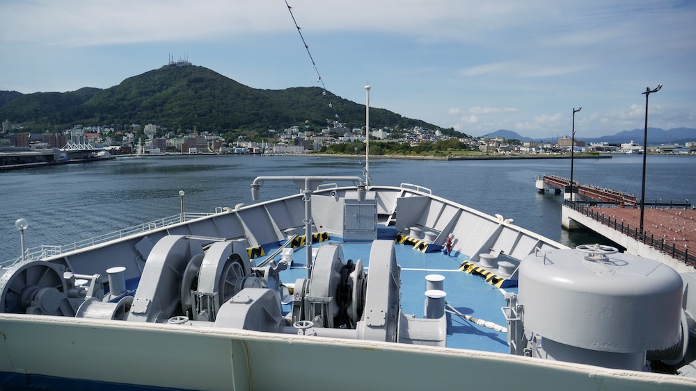 View from the deck of the Hakodate Seikan Ferry Memorial Ship Mashu-maru across Hakodate Bay in Hokkaido, Japan, with Mount Hakodate rising behind the waterfront and harbour.