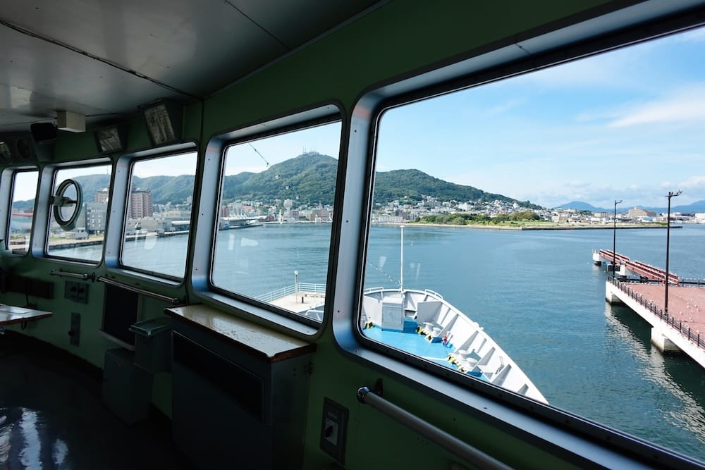 View from inside the bridge of Mashu Maru in Hakodate, Hokkaido, Japan, looking through large windows across the harbour toward the waterfront, city buildings and green slopes beyond.