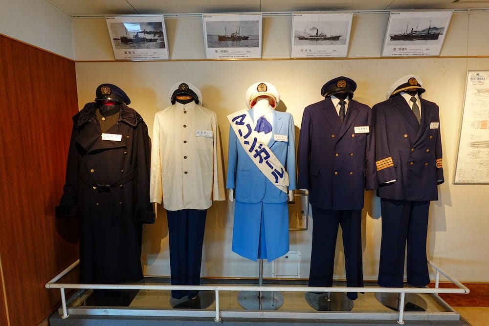 Historic crew uniforms displayed inside Seikan Ferry Memorial Ship Mashu-maru in Hakodate, Hokkaido, Japan, with naval-style jackets, caps and archival ship photographs arranged above the exhibit.