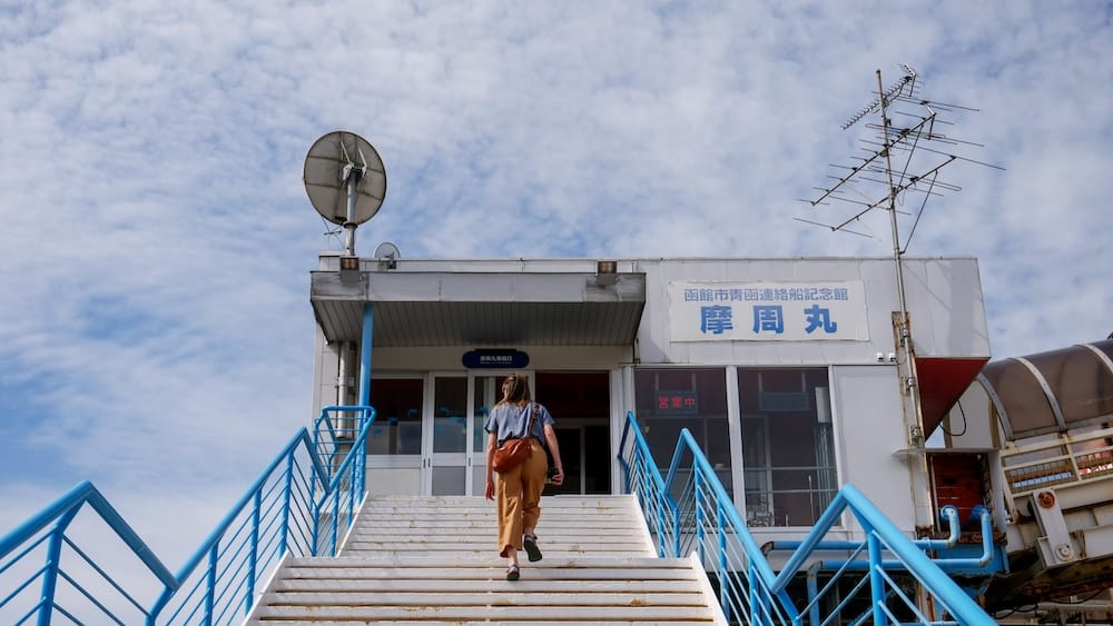 Stairs to Mashu-maru museum ship on the Hakodate waterfront, Hakodate, Hokkaido, Japan – Audrey Bergner Audrey Bergner climbs white stairs with blue railings toward the entrance of Mashu-maru in Hakodate, Hokkaido, Japan, past antennas and a radar dish against a bright, cloudy sky.