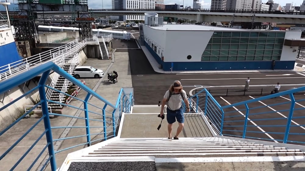 Samuel Jeffery climbs concrete steps with blue railings toward Mashu-maru in Hakodate, Hokkaido, Japan, carrying a camera and wearing a cap and backpack, with a ferry terminal building and parked car below.