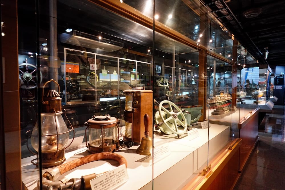 Glass display cases inside the Mashu-maru museum ship in Hakodate, Hokkaido, Japan, showing brass ship lanterns, a bell, a green helm wheel, a flexible speaking tube and model ships in the background.