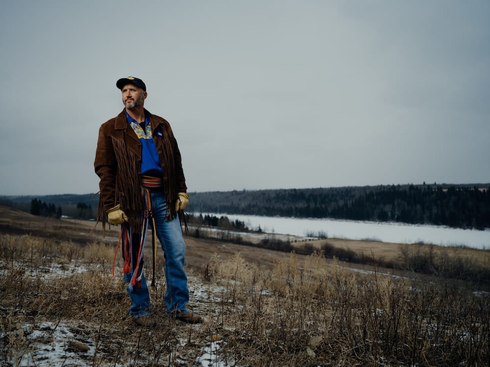A man wearing Métis clothing stands on open grassland at Métis Crossing in Alberta, Canada, with patches of snow, rolling terrain and the North Saskatchewan River valley behind him.