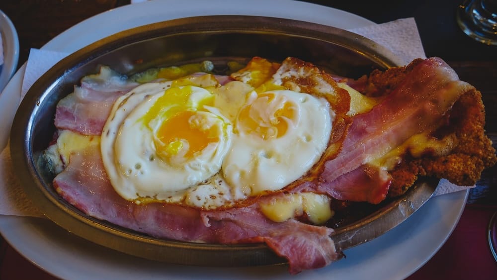 Milanesa a Caballo in El Bolsón Patagonia and Month-by-Month Food Cost Travel Planning Milanesa a caballo with ham, melted cheese and fried eggs in El Bolsón, Patagonia, showing how hearty regional meals become part of the month-by-month travel reality after long buses, cold weather and demanding route days.