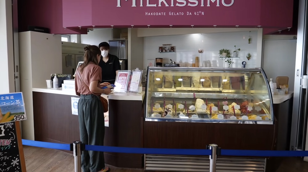 Audrey Bergner hace el pedido en el mostrador de Milkissimo en la Torre Goryokaku, Hakodate, Hokkaido, Japón, junto a una vitrina de vidrio con gelato bajo el cartel Hakodate Gelato da 41°N del local.