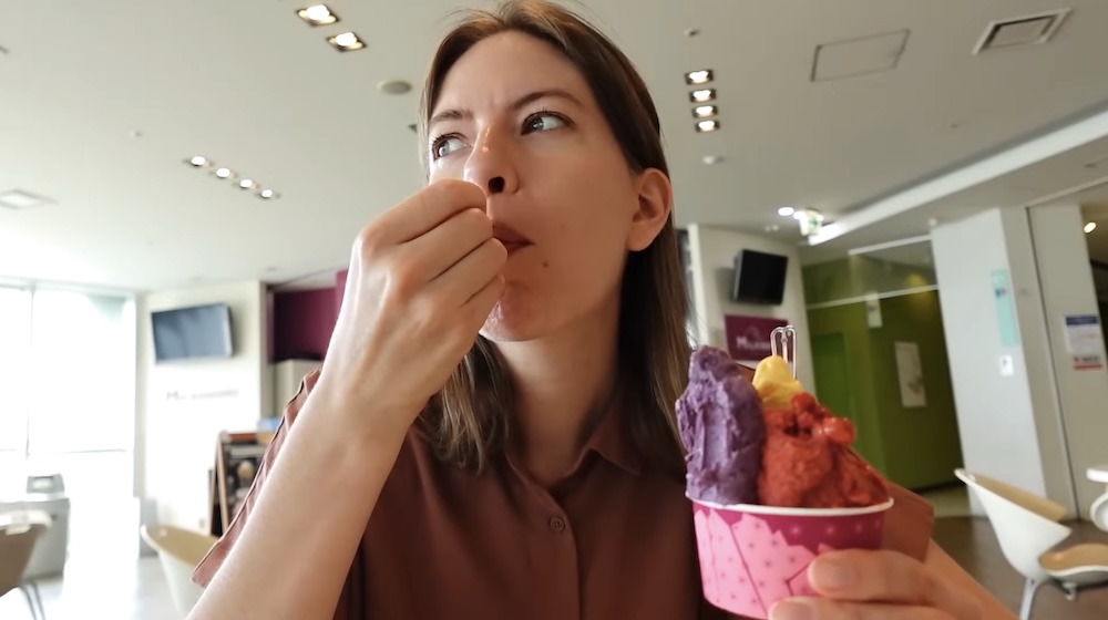 Audrey Bergner come una cucharada de gelato mientras sostiene un vasito rosa con bochas morada, amarilla y roja en el luminoso sector de mesas de Milkissimo dentro de la Torre Goryokaku, Hakodate, Hokkaido, Japón.