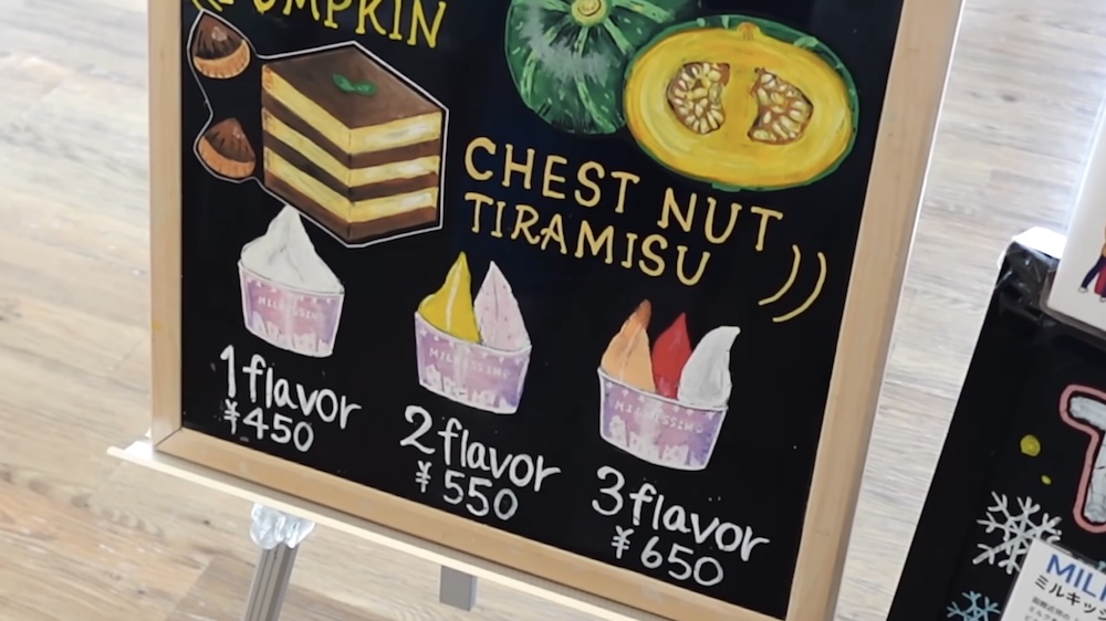 Cartel de pizarrón escrito a mano en Milkissimo dentro de la Torre Goryokaku, Hakodate, Hokkaido, Japón, con dibujos de vasitos de gelato para uno, dos y tres sabores junto a ilustraciones de zapallo y tiramisú de castañas.