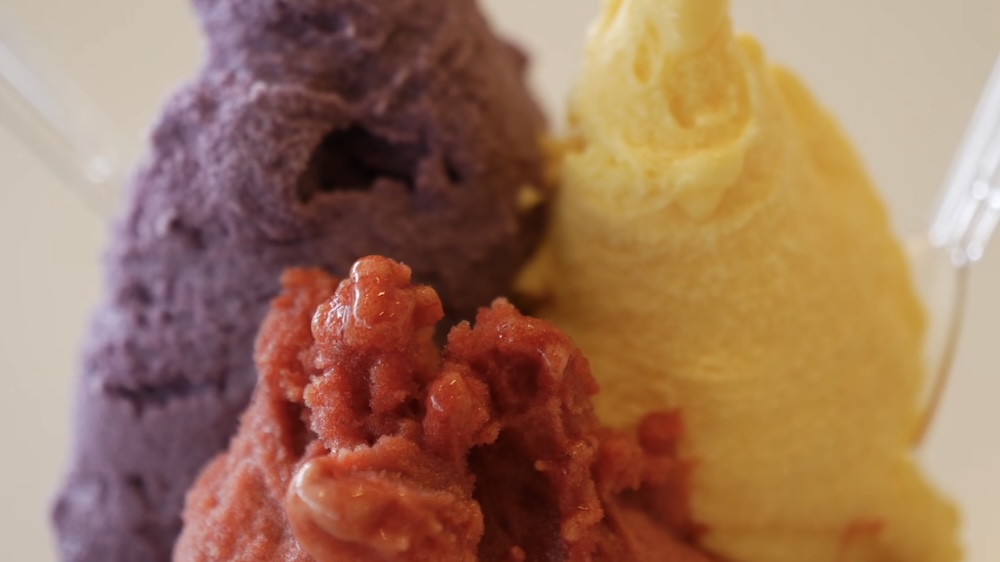 Primer plano macro de tres bochas de gelato lado a lado, batata morada, baya roja y zapallo amarillo, con cucharitas de plástico transparente en los bordes, en Milkissimo dentro de la Torre Goryokaku, Hakodate, Hokkaido, Japón.