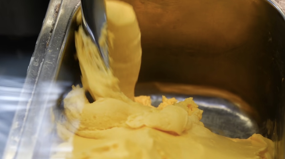Primer plano de una espátula de metal sirviendo gelato suave de zapallo amarillo anaranjado desde una bandeja de acero inoxidable en Milkissimo dentro de la Torre Goryokaku en Hakodate, Hokkaido, Japón.