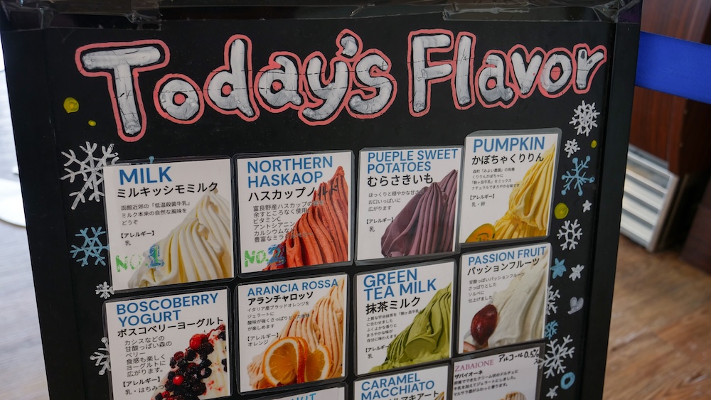 Pizarrón negro Today’s Flavor en Milkissimo dentro de la Torre Goryokaku, Hakodate, Hokkaido, Japón, con fotos de gelato de leche, haskap, batata morada, zapallo, maracuyá y té verde. El cartel de ese día incluía Northern Haskap, que estoy bastante seguro de que era nuestra baya, y zapallo de una granja de Mori, ambos de la línea Hokkaido del local, junto a la batata morada, un sabor de la casa. Cambia todos los días.