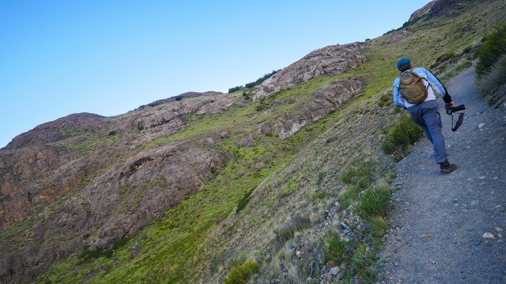 Hiker ascending the Mirador de los Cóndores trail in El Chaltén A hiker (Nomadic Samuel) climbs the dirt trail on the Mirador de los Cóndores hike in El Chaltén, carrying a camera while ascending the rocky Patagonian hillside toward the first panoramic viewpoint above town.