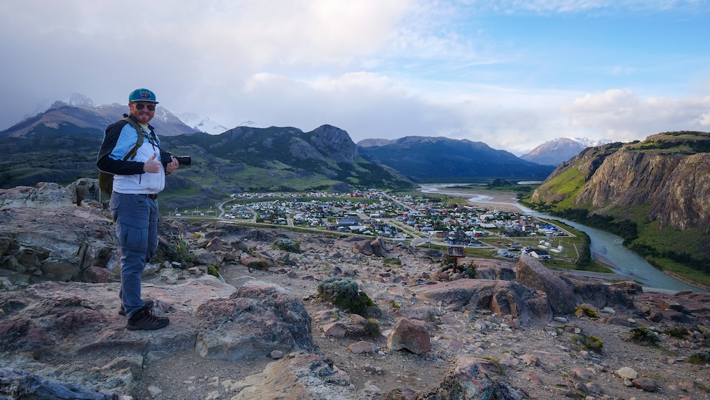 Hiker enjoying Mirador de los Cóndores view over El Chaltén A hiker gives a thumbs up while holding a camera at the Mirador de los Cóndores viewpoint in El Chaltén, overlooking the town, winding river, and rugged Patagonian mountains in the distance.