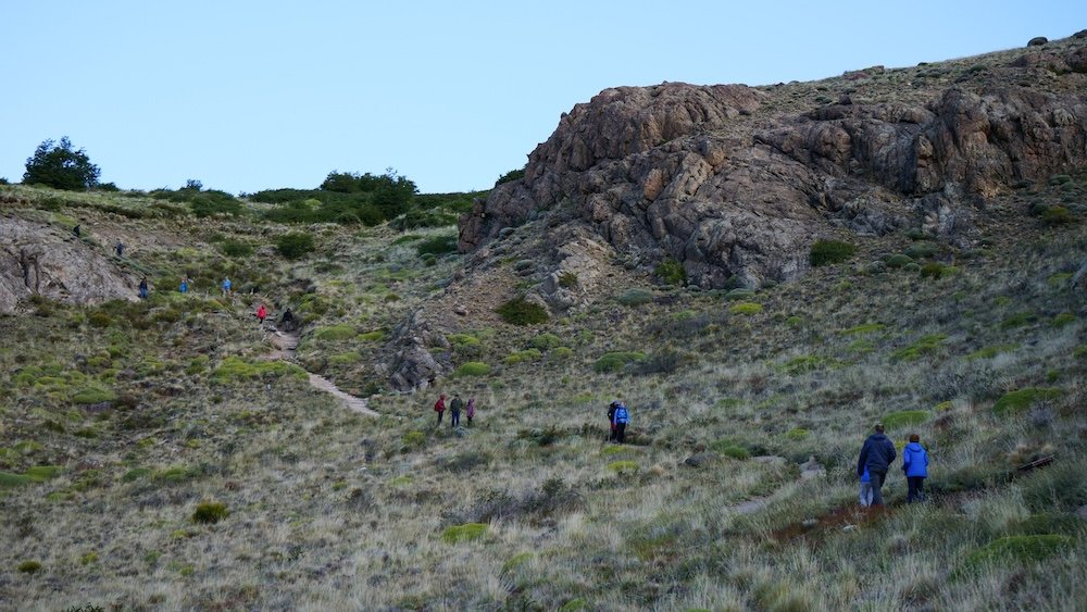 Mirador de los Cóndores, El Chaltén: hikers make their way up a grassy hillside trail with rocky outcrops, starting the classic viewpoint hike that offers sweeping views over town and surrounding Patagonian landscapes inside Los Glaciares National Park.

