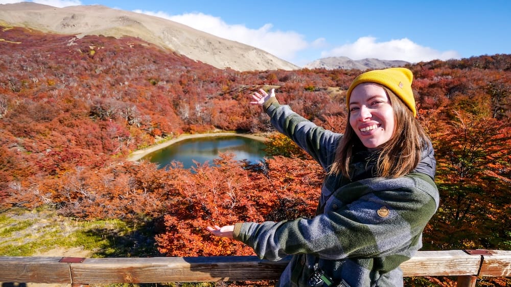 Mirador Laguna Verde Near Lago Gutiérrez, Río Negro, Patagonia, with Audrey Bergner in Autumn Audrey Bergner gestures toward Mirador Laguna Verde near Lago Gutiérrez in Río Negro, Patagonia, Argentina, surrounded by vivid red autumn forest, a small alpine lagoon and pale mountain peaks.