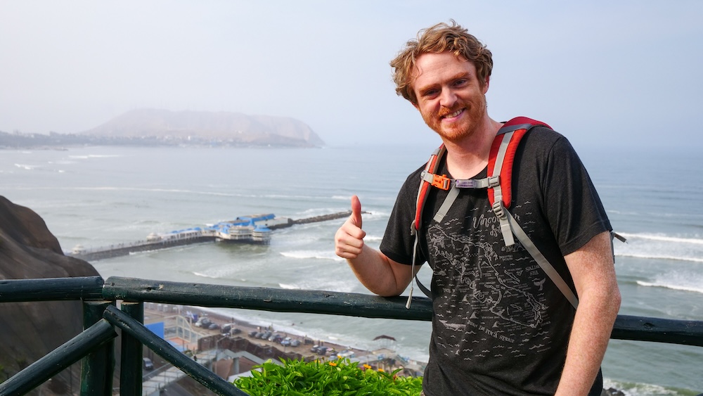 Miraflores, Lima, Peru coastal viewpoint, with Nomadic Samuel wearing a backpack giving a thumbs up on a balcony overlooking the Pacific Ocean, beach promenade, and hazy cliffs on a city travel day.