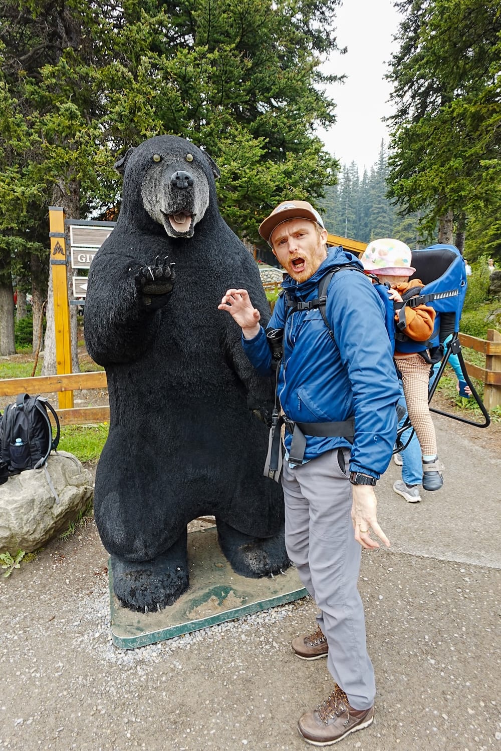 Samuel Jeffery and Aurelia Jeffery with Black Bear Statue at Moraine Lake, Banff National Park, Alberta, Canada Samuel Jeffery poses beside a large black bear statue at Moraine Lake in Banff National Park, Alberta, Canada, while carrying Aurelia Jeffery in a child carrier among evergreen trees near the visitor area.