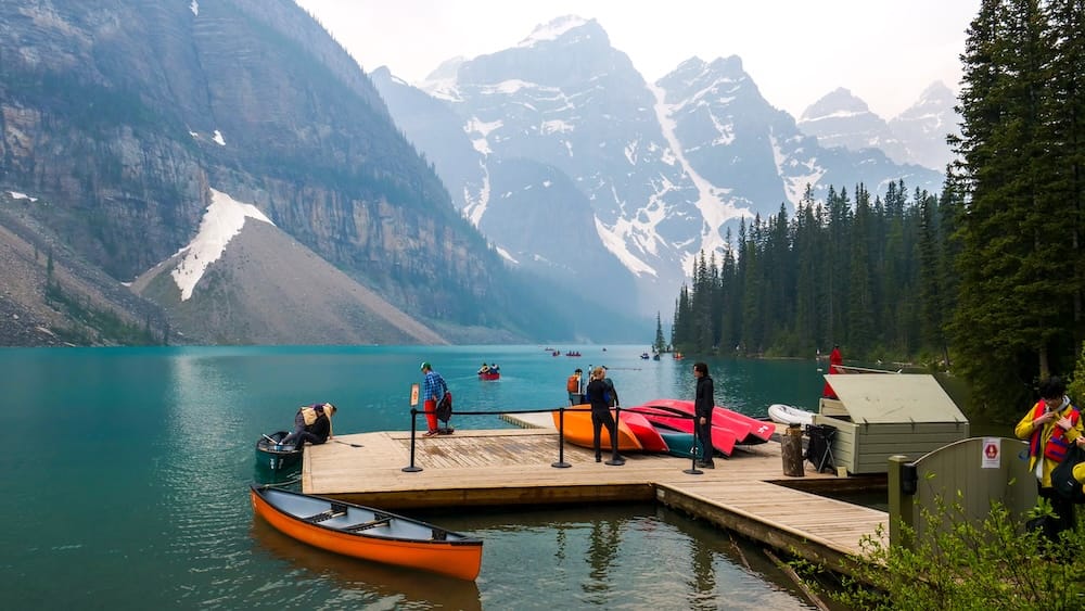 Moraine Lake in Banff National Park, Alberta, Canada, with colourful canoes at the dock, turquoise water, evergreen forest and steep snow-streaked Canadian Rockies rising behind the lake.