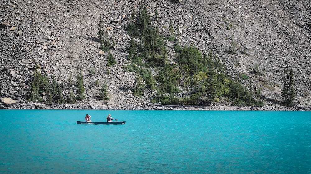 Canoeing on Moraine Lake in Banff National Park, Alberta, Canada, with Turquoise Water and Rocky Mountain Shoreline Two visitors paddle a canoe across the vivid turquoise water of Moraine Lake in Banff National Park, Alberta, Canada, with a steep rocky shoreline and scattered evergreen trees rising behind them.