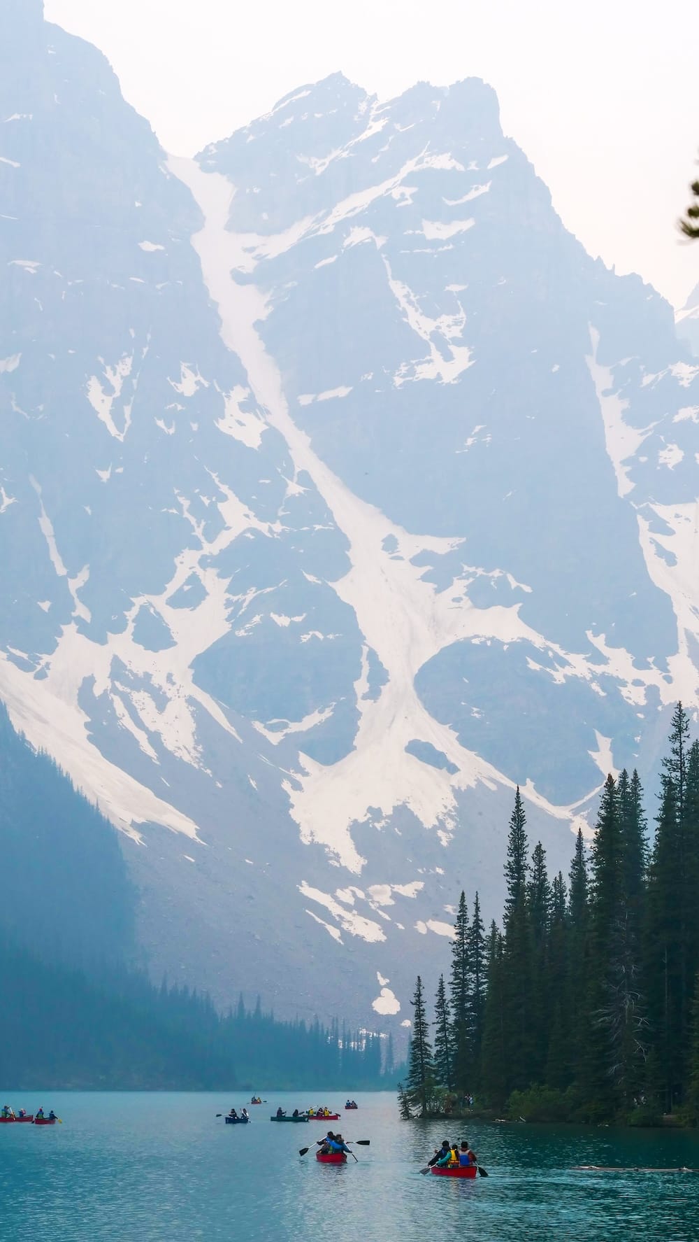 Canoes paddle across the turquoise water of Moraine Lake in Banff National Park, Alberta, Canada, beneath towering snow-streaked mountains and a dense evergreen shoreline.
