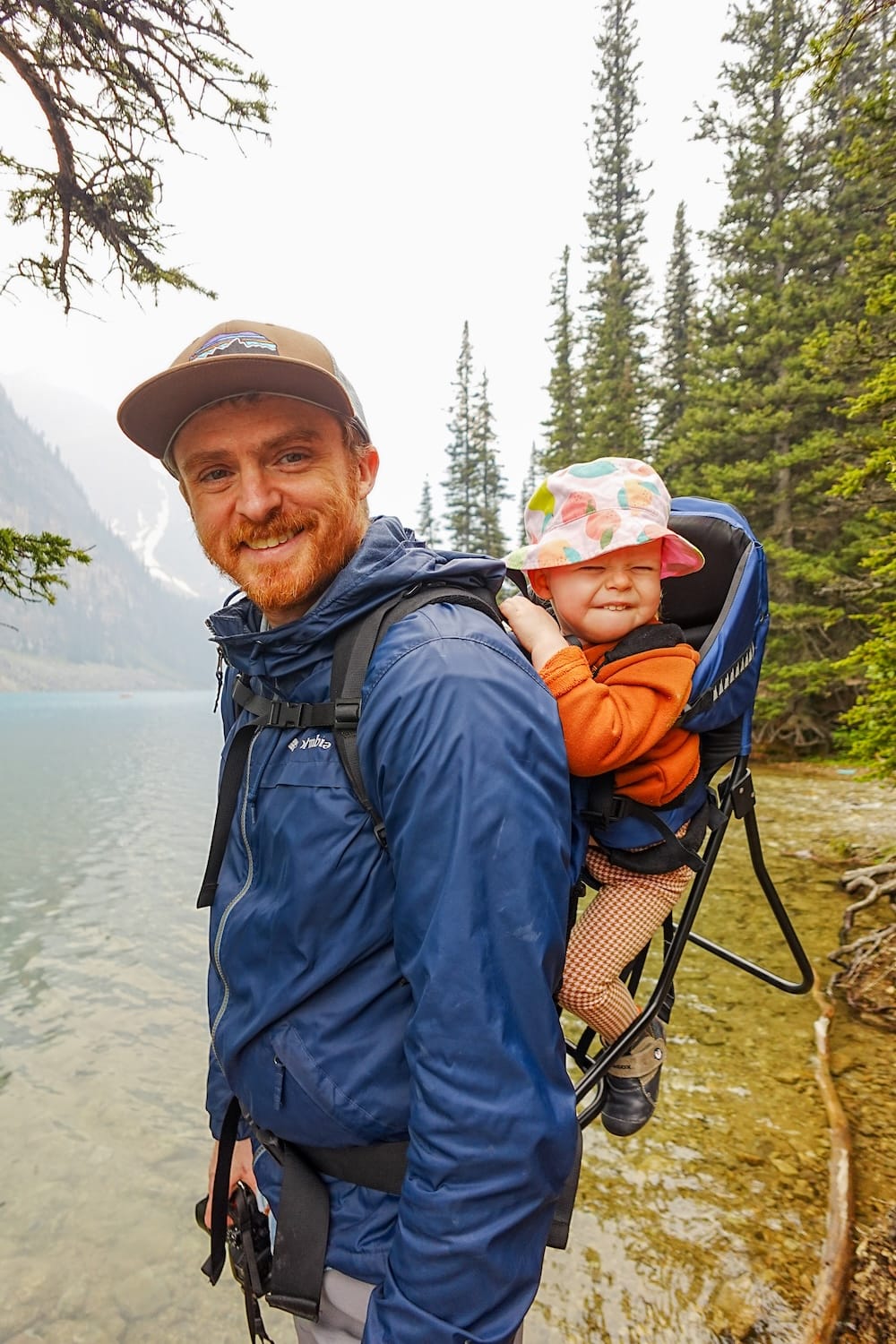 Samuel and Aurelia Jeffery at Moraine Lake, Banff National Park, Alberta, Canada, with child carrier Samuel Jeffery carries Aurelia Jeffery in a child carrier beside the forested shoreline of Moraine Lake in Banff National Park, Alberta, Canada, with mountains fading into the background.